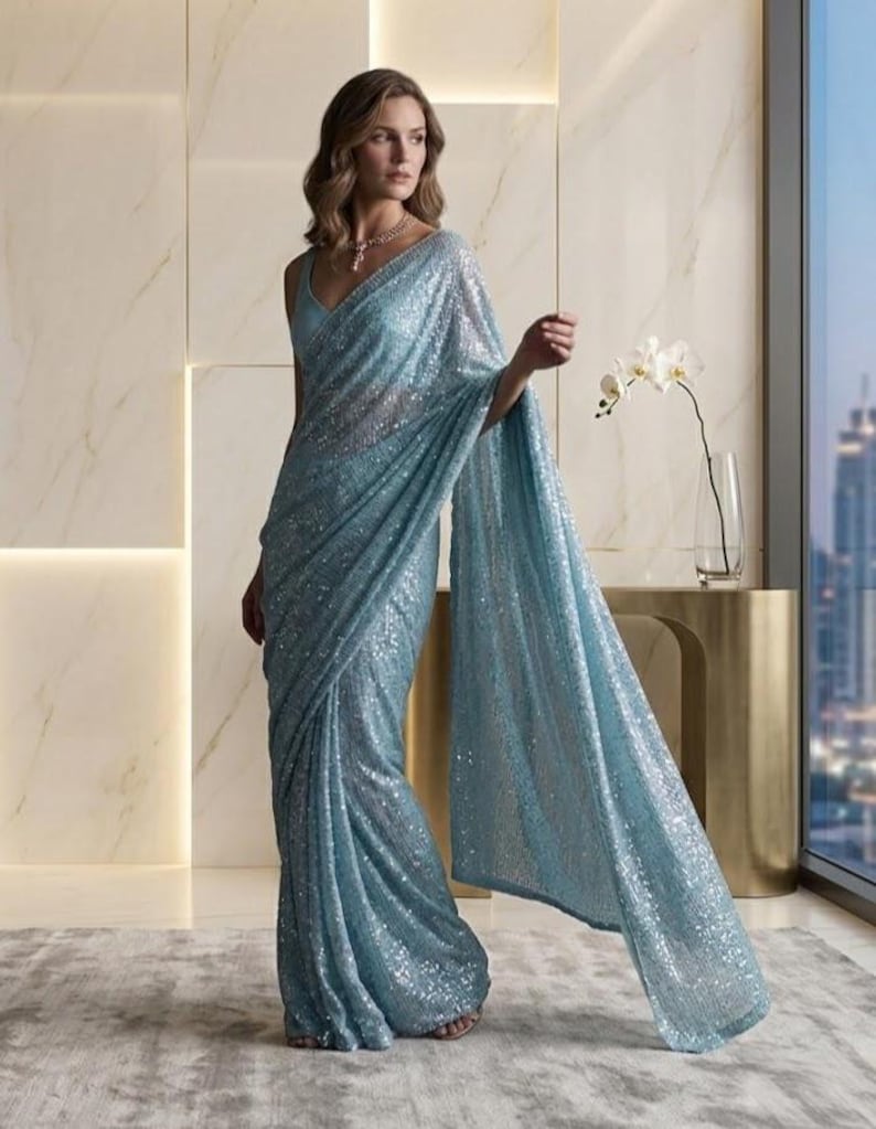 Puede incluir: Un sari azul claro con lentejuelas y un top a juego. El sari cae elegantemente, reflejando la luz con su superficie brillante. El atuendo se complementa con un delicado collar. El fondo presenta un interior moderno con vistas a la ciudad.