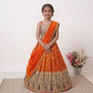 May include: A young girl wearing an orange and gold traditional Indian dress. The dress features a detailed embroidered bodice, a flowing skirt with paisley patterns, and a matching draped shawl. The outfit is vibrant and festive, suitable for special occasions.