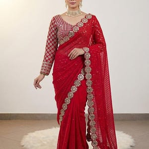 May include: A vibrant red saree with intricate gold embroidery and embellishments. The saree features a detailed border and a matching long-sleeved blouse with similar embellishments. The fabric appears to be sheer, with a delicate pattern throughout.