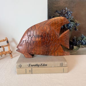 Puede incluir: Una escultura de pez de madera tallada con patrones de escamas detallados. El pez es de color marrón cálido y descansa sobre dos libros. La escultura tiene un diseño detallado de aleta y cola.