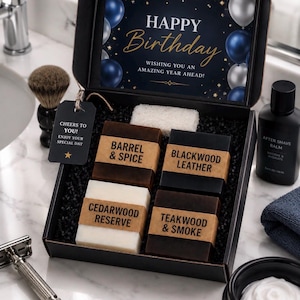 以下が含まれることがあります： Barrel & Spice、Blackwood Leather、Cedarwood Reserve、Teakwood & Smokeとラベル付けされた4つの長方形の石鹸が入った黒いギフトボックス。蓋の内側にはHappy Birthdayの文字と青と銀の風船が描かれています。タグにはCheers to Youと書かれています。