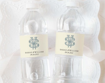 May include: Two clear plastic water bottles with cream-colored labels. The labels feature a blue monogram with the letters "S" and "W" and the text "Sarah & William 09.24.2022". The bottles are on a white lace doily.