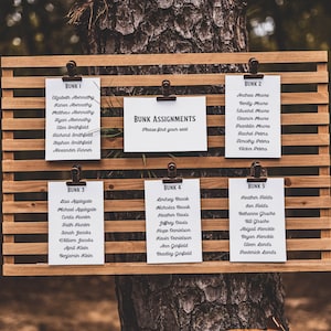May include: A wooden sign with a grid pattern and clips holding white cards with black text. The cards list names under the headings "Bunk 1", "Bunk 2", "Bunk 3", "Bunk 4", and "Bunk 5". A separate card reads "Bunk Assignments, Please find your seat."