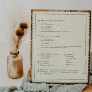 May include: A gold-framed sign with a white background and black text lists the drinks and cocktails available at a wedding. The sign is titled "The Mixology of Sarah & William's Wedding". The drinks include "The Catcher in the Rye", "Scotch Fizz-Gerald", "Cabernet Sauvignon", "Pinot Grigio", "Prosecco", "Bud Light", "Keystones", "Michelob Ultra", "Pineapple", "Orange", "Cranberry", "Coke", "Gingerale", "Rootbeer", "Seagrams", "Gray Goose", "Kahula", "Contreau", "Amontillado Sherry", "Single Malt Scotch", and "Grand Marnier".