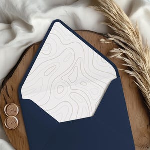 May include: A navy blue envelope with a white interior featuring a gray topographic design. The envelope is open, revealing the patterned interior. Two gold wax seals with initials are visible, along with dried ornamental grass.