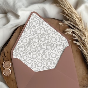 May include: A dusty rose envelope with a geometric patterned interior. The envelope is open, revealing a gray and white sunburst design. Two gold wax seals with initials are visible, along with dried ornamental grass.