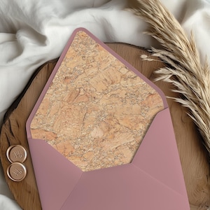 May include: A dusty rose envelope with a cork interior, open to reveal the textured pattern. The envelope sits on a wooden surface, with two gold wax seals and dried ornamental grass. The background is a cream-colored fabric.