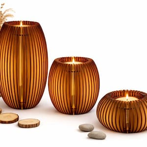 Puede incluir: Tres candelabros de madera con velas encendidas, de diferentes tamaños y formas, están colocados sobre una superficie blanca. Los candelabros tienen un brillo ámbar cálido. Rodajas de madera decorativas y pequeñas piedras están dispersas.