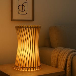 Pode incluir: Uma luminária de mesa de madeira com um design único. A luminária apresenta ripas de madeira verticais que se curvam em torno da fonte de luz, criando um brilho quente. A luminária está sobre uma mesa lateral de madeira, com uma obra de arte emoldurada ao fundo.