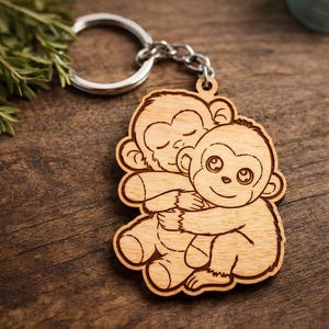 May include: Wooden keychain featuring two hugging cartoon monkeys. The keychain has a silver metal ring and chain. The monkeys are light brown with etched details, one with closed eyes and the other with open eyes and a smile.