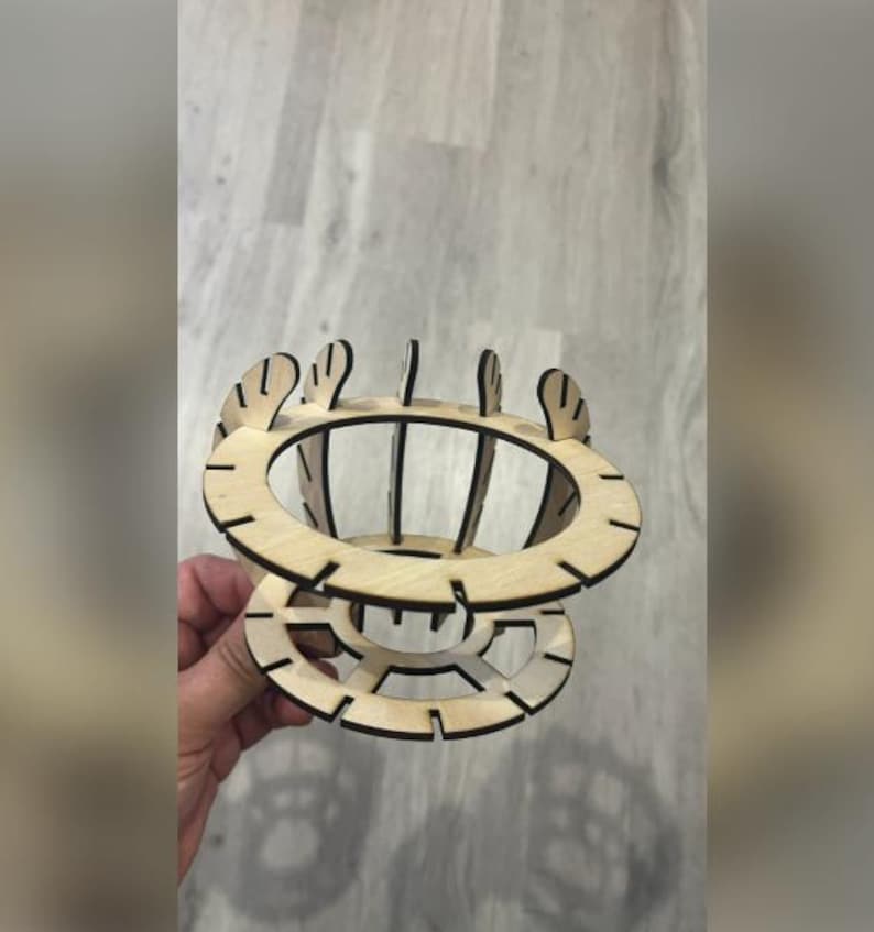 Puede incluir: Un objeto decorativo de madera cortado con l&aacute;ser con un dise&ntilde;o circular. El objeto presenta m&uacute;ltiples capas y soportes verticales, creando una estructura abierta en forma de jaula. El color natural de la madera es visible.