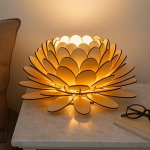 Puede incluir: Una l&aacute;mpara de madera con forma de flor y p&eacute;talos superpuestos, que emite un brillo c&aacute;lido. La l&aacute;mpara est&aacute; sobre una mesita de noche con tapa de m&aacute;rmol blanco, junto a un par de gafas y un libro. La l&aacute;mpara mide unos 30 cm de di&aacute;metro.
