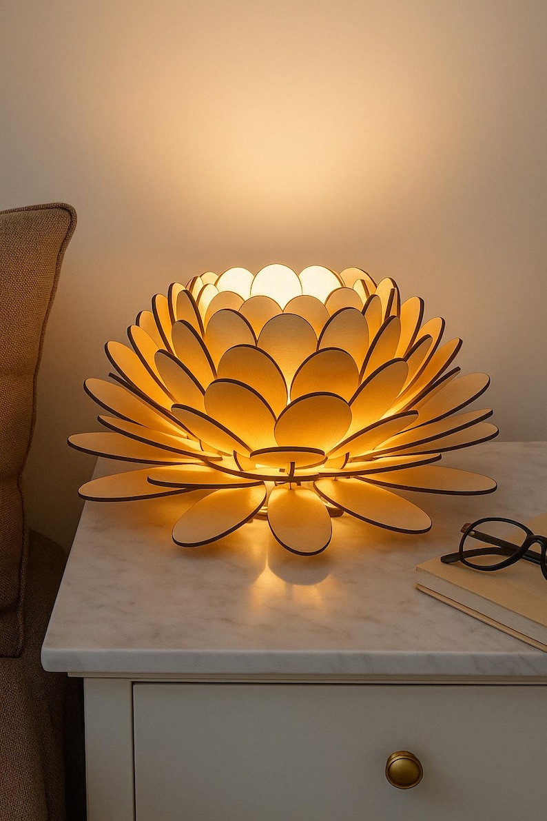 Puede incluir: Una l&aacute;mpara de madera con forma de flor y p&eacute;talos superpuestos, que emite un brillo c&aacute;lido. La l&aacute;mpara est&aacute; sobre una mesita de noche con tapa de m&aacute;rmol blanco, junto a un par de gafas y un libro. La l&aacute;mpara mide unos 30 cm de di&aacute;metro.