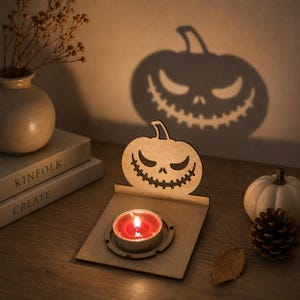 May include: A wooden Halloween pumpkin tea light holder with a carved jack-o'-lantern face. The candle is lit, casting a shadow of the pumpkin face on the wall. A small white pumpkin and pine cone are also visible.