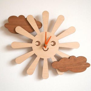 May include: A wooden wall clock in the shape of a smiling sun with orange clock hands. Two brown wooden clouds are positioned on either side of the sun. The clock is made of light-colored wood.