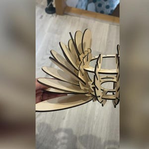 Puede incluir: Un objeto decorativo de madera cortado con l&aacute;ser, con un dise&ntilde;o en capas en forma de abanico. El objeto est&aacute; construido con madera de color claro con bordes marr&oacute;n oscuro, creando un contraste. El dise&ntilde;o parece una flor o un p&aacute;jaro estilizado.