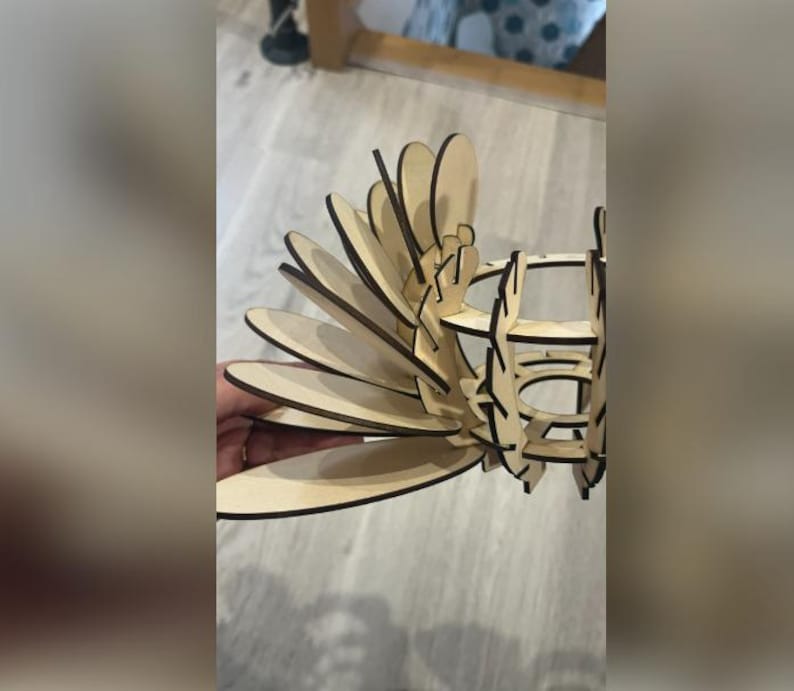 Puede incluir: Un objeto decorativo de madera cortado con l&aacute;ser, con un dise&ntilde;o en capas en forma de abanico. El objeto est&aacute; construido con madera de color claro con bordes marr&oacute;n oscuro, creando un contraste. El dise&ntilde;o parece una flor o un p&aacute;jaro estilizado.