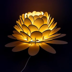 Puede incluir: L&aacute;mpara decorativa con forma de flor, con elementos en forma de p&eacute;talos superpuestos. La l&aacute;mpara emite un brillo amarillo c&aacute;lido, creando un ambiente suave y acogedor. El dise&ntilde;o presenta un cable blanco para la alimentaci&oacute;n, sobre un fondo oscuro.