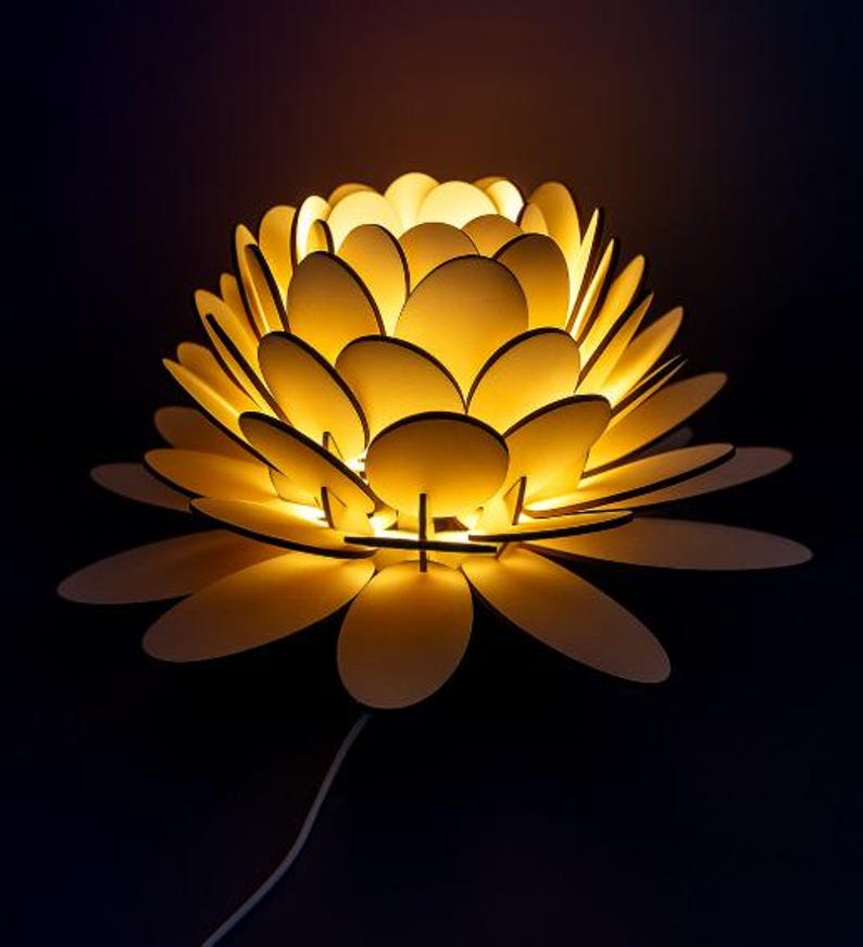 Puede incluir: L&aacute;mpara decorativa con forma de flor, con elementos en forma de p&eacute;talos superpuestos. La l&aacute;mpara emite un brillo amarillo c&aacute;lido, creando un ambiente suave y acogedor. El dise&ntilde;o presenta un cable blanco para la alimentaci&oacute;n, sobre un fondo oscuro.