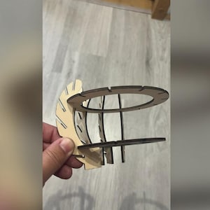 Puede incluir: Objeto decorativo de madera cortada con l&aacute;ser, sostenido por una mano. El objeto tiene una parte superior e inferior circulares, conectadas por soportes verticales. Una pieza curva en forma de abanico est&aacute; unida al lateral. La madera es de color claro natural.