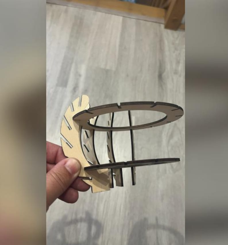 Puede incluir: Objeto decorativo de madera cortada con l&aacute;ser, sostenido por una mano. El objeto tiene una parte superior e inferior circulares, conectadas por soportes verticales. Una pieza curva en forma de abanico est&aacute; unida al lateral. La madera es de color claro natural.