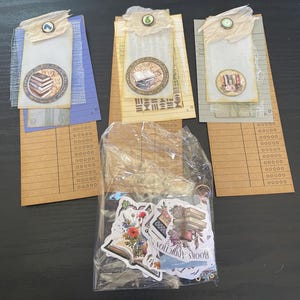 May include: A collection of handmade bookmarks and stickers. The bookmarks feature vintage-style designs with book illustrations and decorative elements. The stickers, in a clear bag, have floral and book graphics. The text "BOOKS ADDICTION" is visible on the stickers.