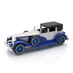 May include: A miniature classic car model in silver, blue, and black. The car features a convertible top, blue wheels with white-walled tires, and detailed chrome accents. A vintage-style collectible.