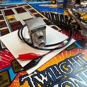 May include: A silver toy camera with the words "Rock n Roll" printed on the front. The camera is connected to a black wire with red and black alligator clips. The camera is sitting on a white piece of paper on top of a pinball machine with a blue and yellow design.