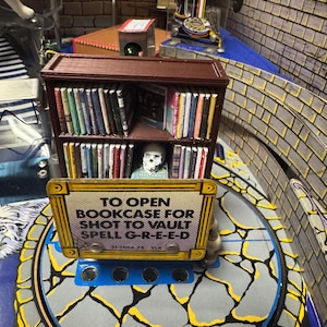 May include: A brown wooden bookcase with a sign that reads "To open bookcase for shot to vault spell G-R-E-E-D". The bookcase is filled with books and has a skull on the top shelf. The sign is yellow with black lettering and has a gold border.