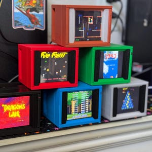 Könnte beinhalten: Sechs Miniatur-Arcade-Spielkonsolen in verschiedenen Farben, die jeweils ein klassisches Spiel anzeigen. Die Konsolen sind rot, orange, grün, blau, schwarz und grau, mit Spieltiteln wie "Free Fight" und "Dragon's Lair" sichtbar.