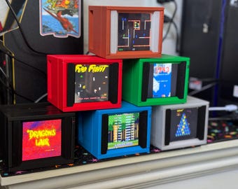 Puede incluir: Una colección de seis mini consolas de juegos arcade en varios colores, cada una mostrando un juego clásico diferente. Las consolas son rojas, naranjas, verdes, azules, negras y grises, con títulos de juegos como "Free Fight" y "Dragon's Lair" visibles.