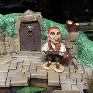 Op de afbeelding: Een miniatuurbeeldje van een goblinachtig personage dat een houten voorwerp vasthoudt, voor een stenen gebouw met twee houten deuren. De figuur heeft rood haar en draagt een wit overhemd en een bruin vest. De scène speelt zich af op een stenen oppervlak.