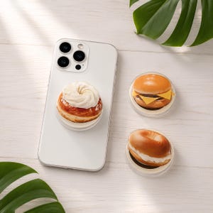 May include: A white smartphone with a circular phone grip featuring a cream-topped pastry design attached to the back. Two additional circular phone grips are nearby, one shaped like a cheeseburger and the other like a cream-filled bagel.