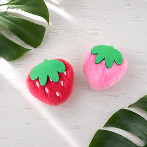 May include: Two plush strawberry-shaped items, one in bright red with white embroidered seed details and one in soft pink, both featuring a green felt leaf topper. These fuzzy, rounded objects are positioned on a white wooden surface.