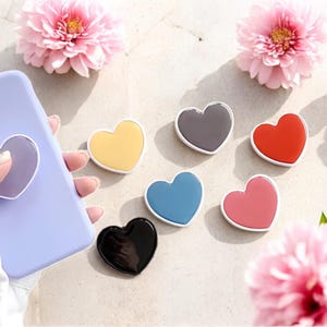 May include: A variety of heart-shaped phone grips in multiple colors, including yellow, gray, red, blue, pink, and black, with white borders. A lavender phone case with a matching heart grip is held in the image. Pink flowers are in the background.