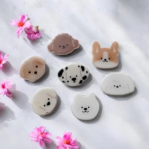 Op de afbeelding: Een verzameling van zeven decoratieve hondenpinnen in verschillende rassen, waaronder een Dalmatische hond en een Corgi. De pinnen zijn in bruin, wit en zwart, met schattige gezichtskenmerken. Roze bloemen zijn verspreid.