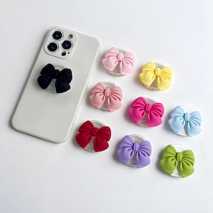 Peut inclure: Un smartphone blanc avec un support de téléphone en forme de nœud noir attaché. Plusieurs supports de téléphone en forme de nœud colorés en rose, jaune, rouge, violet, vert et bleu sont éparpillés autour du téléphone. Les supports ont une base transparente.