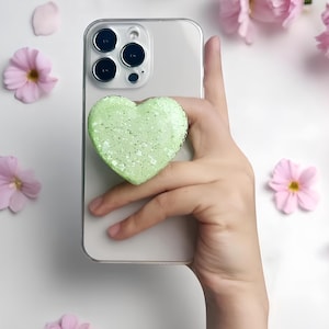 May include: A light green heart-shaped phone grip with glitter is attached to a silver phone. The phone is held by a hand, surrounded by pink flowers and petals on a white surface. The grip adds a decorative touch.