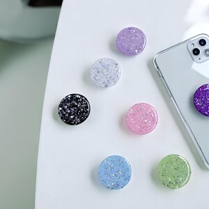 Puede incluir: Una colección de agarres para teléfono de colores con detalles de purpurina. Los agarres son de varios colores, incluyendo morado, rosa, azul, verde, plateado y negro. Un agarre está adherido a un smartphone plateado. Los agarres son redondos y tienen un acabado brillante.