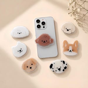 Puede incluir: Un teléfono inteligente gris claro con un soporte para teléfono en forma de caniche marrón en la parte posterior, rodeado de seis soportes adicionales con temática de perros, incluyendo un dálmata, un corgi y varios perros blancos y canela.