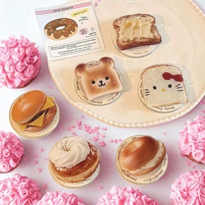 May include: A collection of food-themed decorative items, including a burger, donuts, toast with butter, and toast with a bear and cat design. The items are arranged on a white plate and small clear coasters. Pink floral decorations surround the plate.