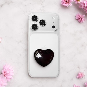 Peut inclure: Un étui de téléphone transparent avec un support de téléphone en forme de cœur noir est centré sur une surface blanche. Des fleurs roses sont disposées autour du téléphone. Le téléphone a trois objectifs.