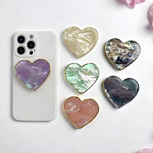 Peut inclure: Un smartphone blanc avec un support de téléphone en forme de cœur violet est présenté avec cinq autres supports en forme de cœur. Chaque support a une bordure dorée et une finition nacrée et irisée. Les supports sont de différentes couleurs et sont disposés sur une surface blanche.