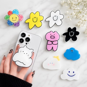 May include: A collection of phone grips featuring various shapes and designs, including a multi-coloured flower, yellow and white stars, a pink pig, a black star, and several blue and white clouds with smiling faces, plus a white cat phone grip.