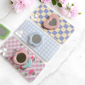 May include: An assortment of phone cases with decorative mirrors. The cases feature various patterns, including a pink and white checkered design, a blue and white gingham pattern, and a yellow and purple checkerboard. The mirrors are heart, flower, and square shaped.