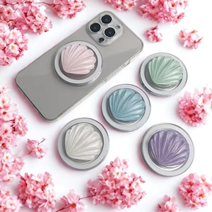 May include: A collection of seashell-shaped phone grips in various pastel colors, including pink, blue, green, white, and purple. One grip is attached to a silver phone. The grips are arranged on a white surface, surrounded by pink flowers.