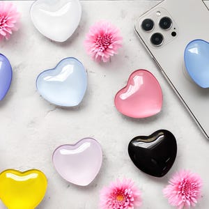 Puede incluir: Una colección de agarres para teléfono con forma de corazón en varios colores, incluyendo blanco, azul, rosa, amarillo y negro. Flores rosas están dispersas alrededor de los agarres. Un agarre azul está adherido a un smartphone plateado.