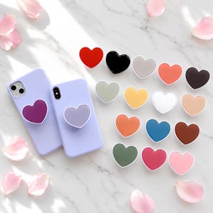 Peut inclure: Deux coques de smartphone violet clair, chacune dotée d'un support de téléphone en forme de cœur. Un support est violet foncé, l'autre est lavande clair. À côté se trouvent seize supports en forme de cœur de couleurs unies variées : rouge, noir, gris, orange, marron, jaune, blanc, beige, bleu, vert, rose et magenta.