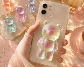 以下が含まれることがあります： 背面に立体的な虹色のクマ型グリップが付いた透明なスマートフォンケース。クマは半透明でキラキラした仕上げが施されており、紫、緑、金の色合いが混ざり合っています。周囲には同様のクマのフィギュアがいくつか配置されています。