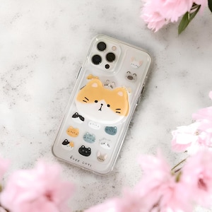 以下が含まれることがあります： オレンジと白の漫画の猫のデザインの透明な携帯電話ケース。ケースには、複数の小さな猫のイラストと「free cats」の文字が描かれています。ピンクの花が携帯電話を囲んでいます。